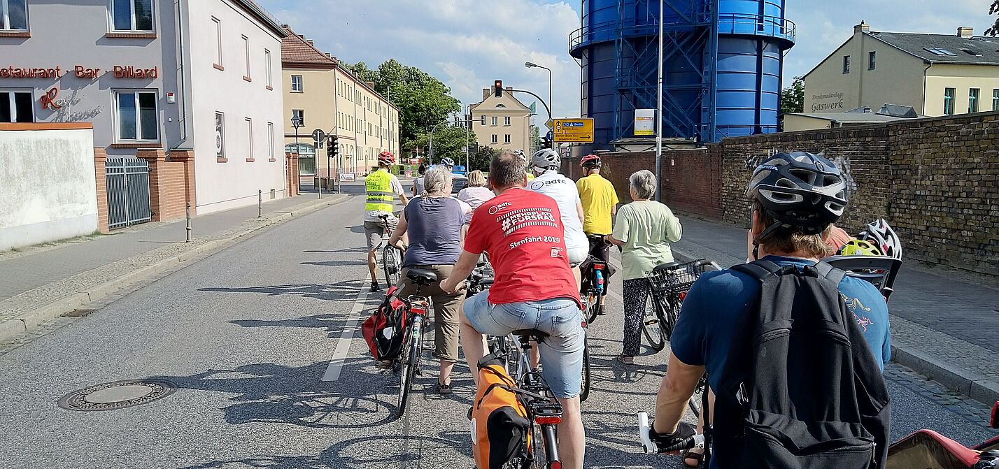 Fahrraddemo in Bernau Das Bild zeigt eine Gruppe von Radfahrern auf einer städtischen Straße. Sie stehen hintereinander auf ihren Fahrrädern und tragen teils Helme sowie Rucksäcke. Links sind Wohnhäuser, rechts eine Backsteinmauer und der Bernauer Gaskessel (ein großes, blaues, rundes Industriegebäude) zu sehen. Das Wetter ist sonnig und freundlich.